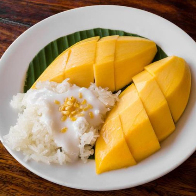 Mango sticky rice with coconut milk and toasted mung beans on a white plate with a banana leaf.